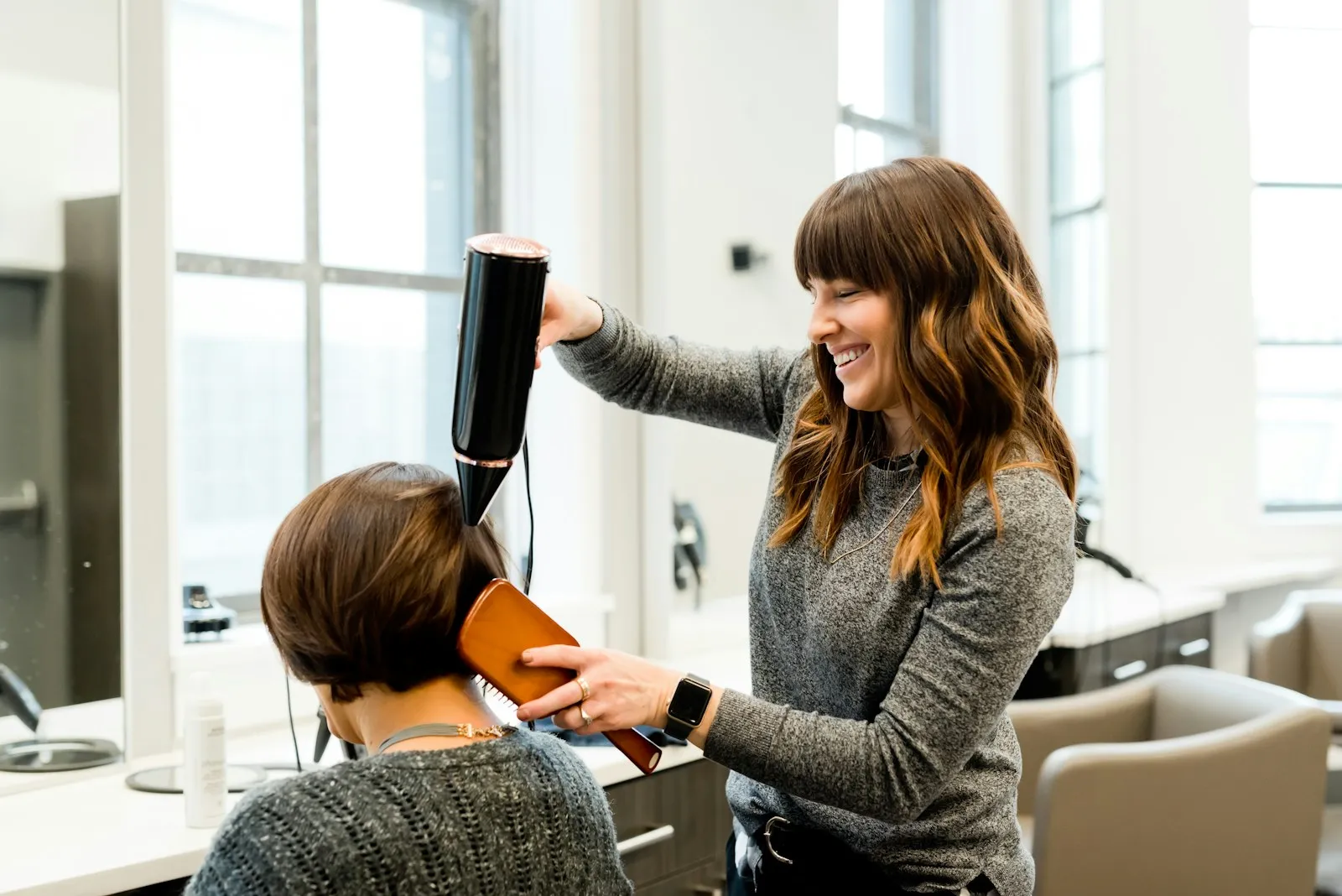 A stylist smiling as she blow-dries a client's hair by a window